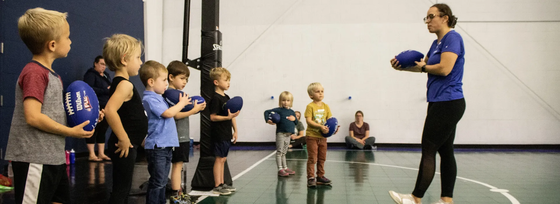 A YMCA team member instructs young children holding small, blue footballs on an indoor court