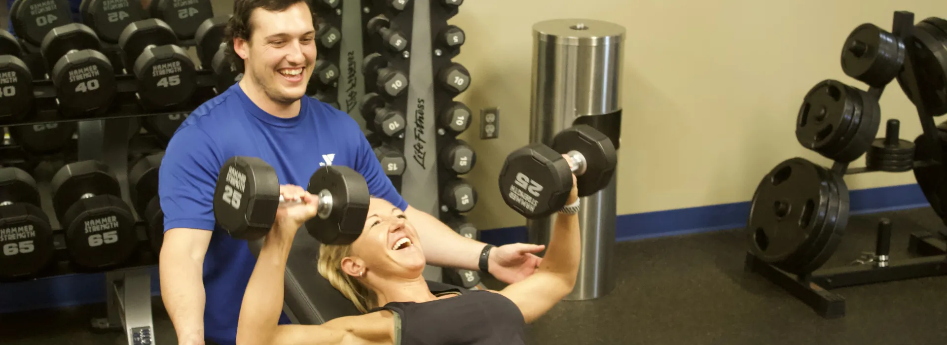 A physical trainer guides a student as they lift 25lb weights.