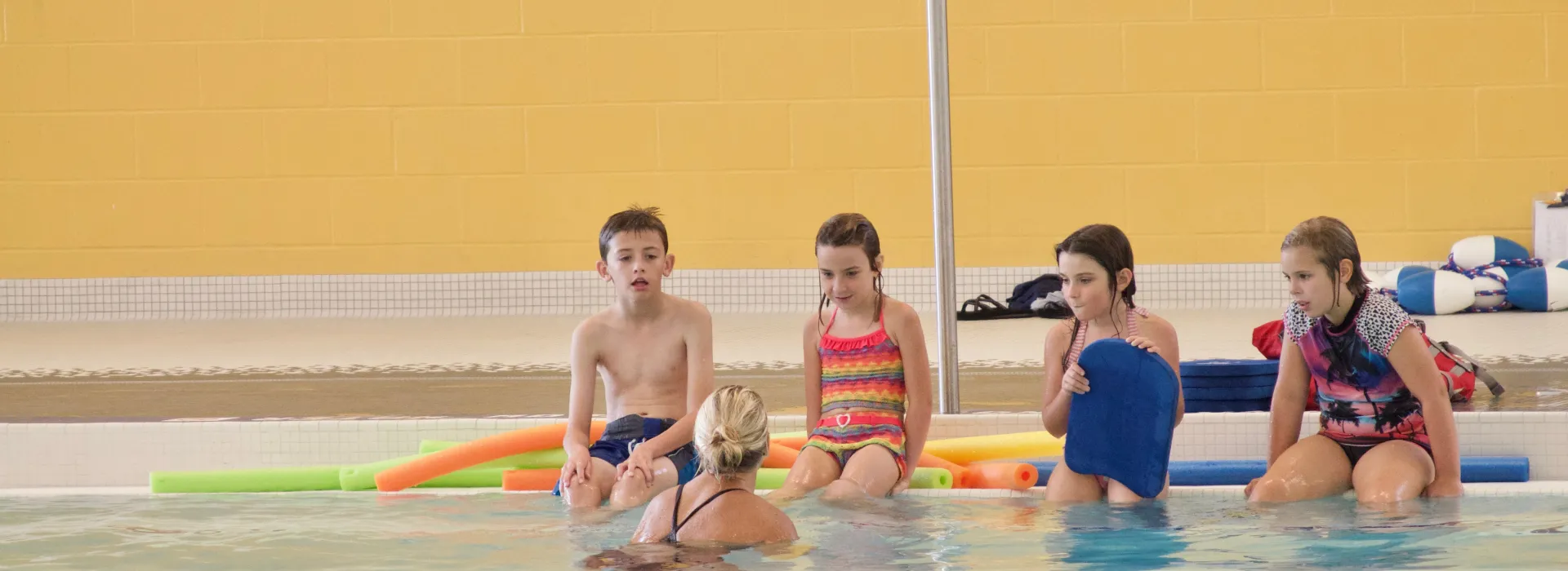 Young children sit on the pool edge, listening to an instructor demonstrating in the water.