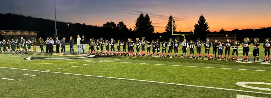 Wide view of young football players lined up on the field as a sun sets in the background.