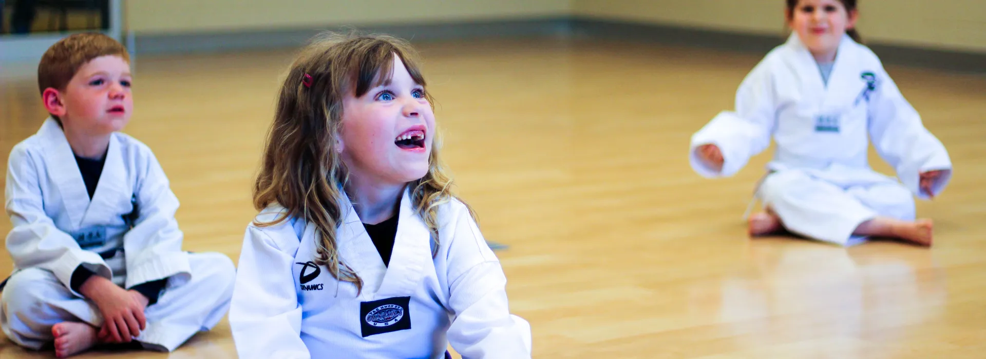 Young Taekwondo students sit on the floor, observing their teacher.