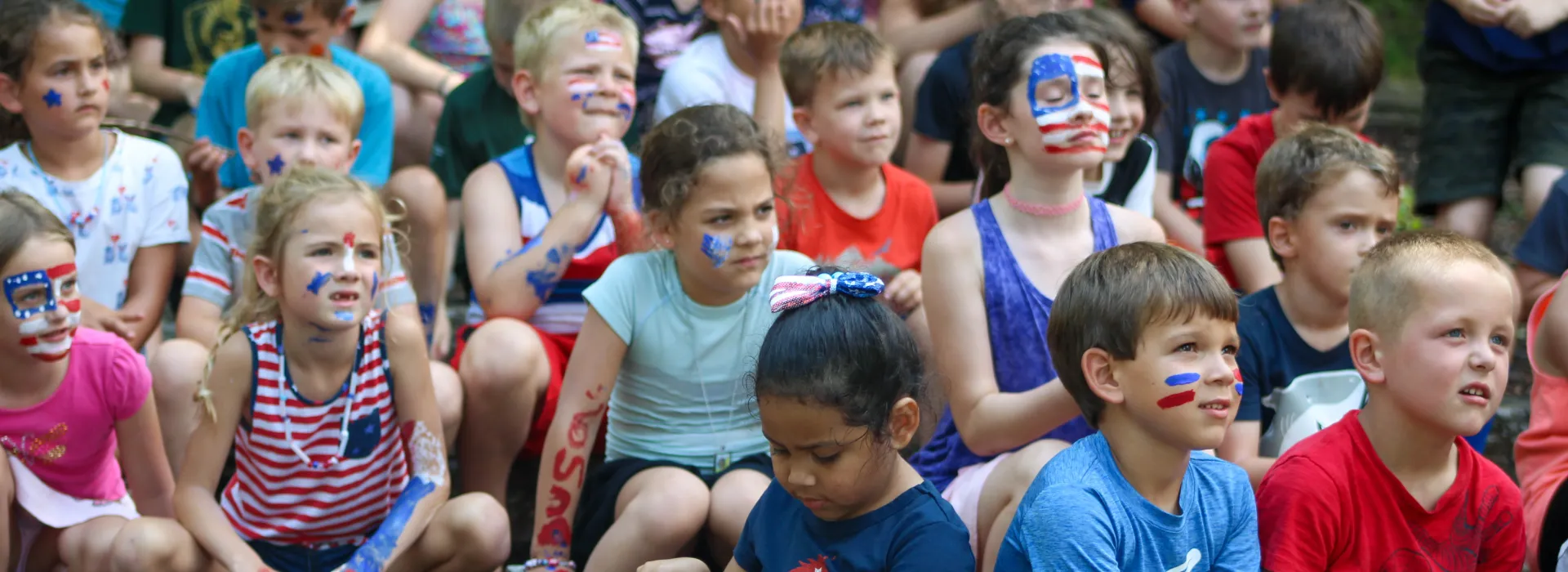 Young children in red, white and blue face paint sit outdoors, listening to a presenter