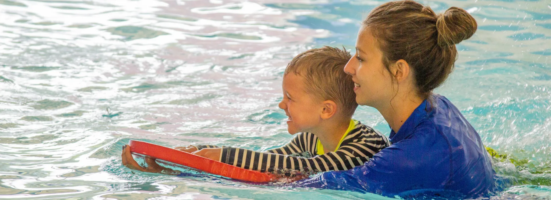 A YMCA instructor helps a young student swim using a kickboard.