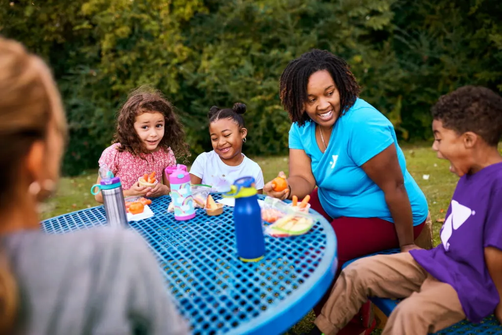 Y Team Member sits at table with children, eating lunch and laughing.