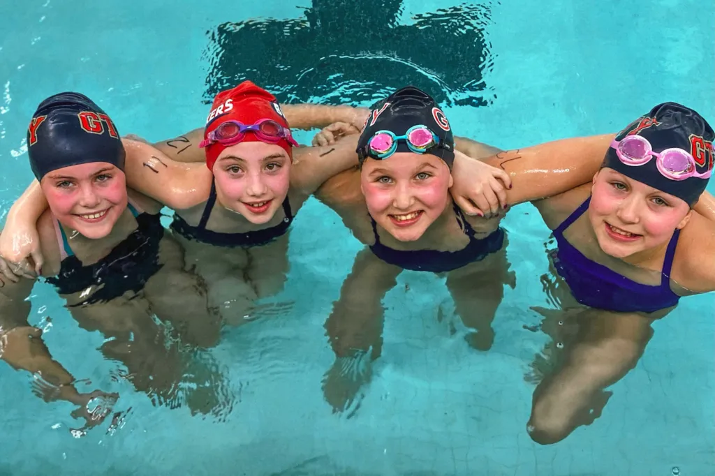 Four swimmers in the pool lock arms and look up, smiling at the camera