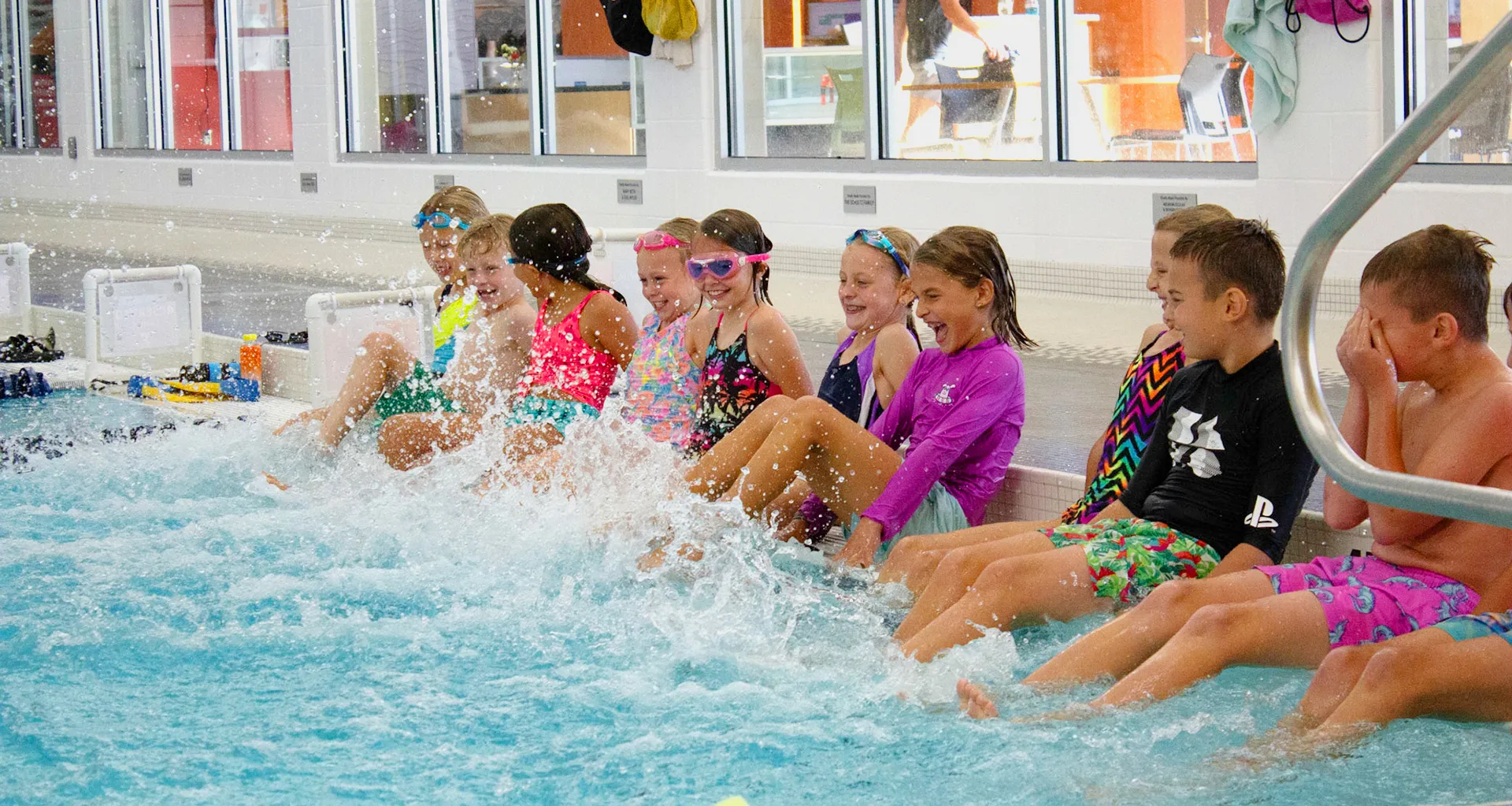 Children in swimwear sit at the pool edge and kick their feet, laughing as water splashes