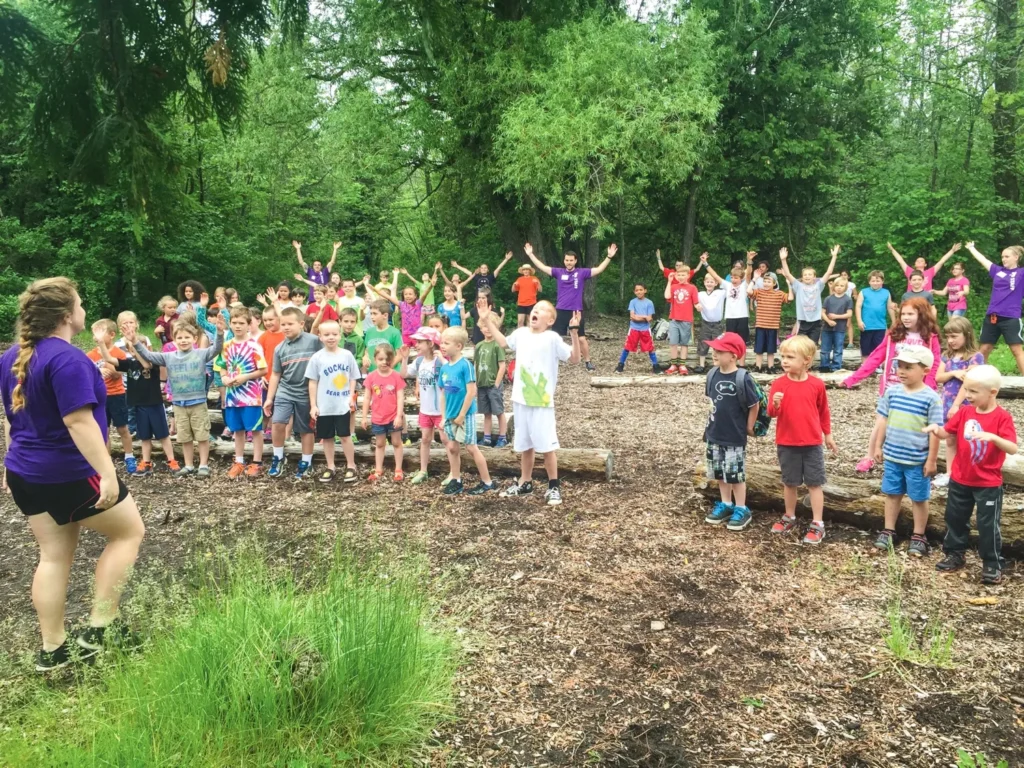 Campers at an outdoor assembly stand and cheer with a counselor