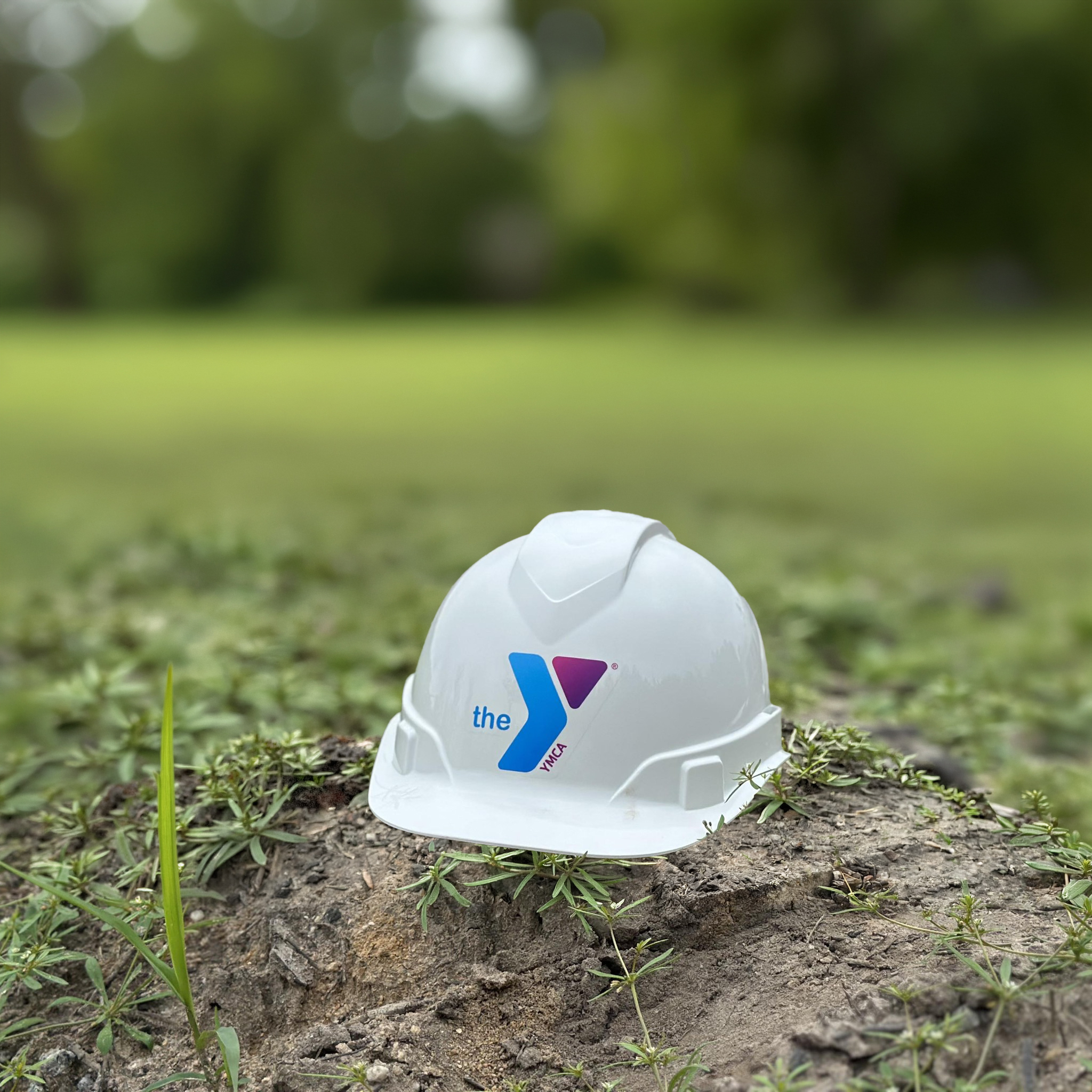 White construction hardhat with a YMCA logo on the front, sitting on a pile of dirt in a grassy green area